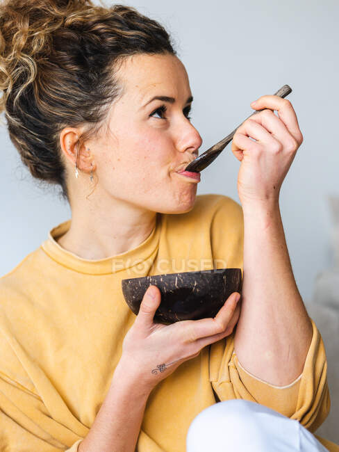 Jovem loira alegre mulher em roupas casuais comer comida saudável de coco natural tigela na sala de luz — Fotografia de Stock