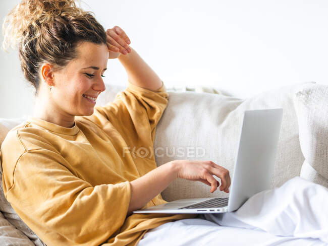 Positif jeune femme avec les cheveux bouclés en vêtements décontractés assis sur un canapé confortable et netbook de navigation dans l'appartement léger en journée — Photo de stock