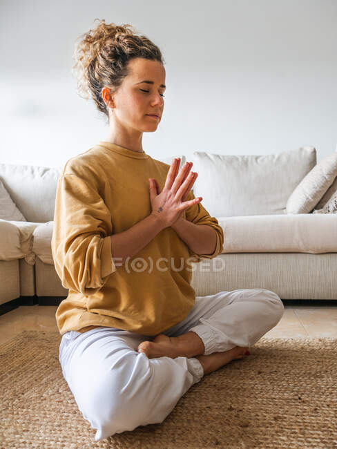 Vista laterale di donna calma in abbigliamento sportivo seduta sul pavimento in posa loto con le mani di preghiera durante la meditazione a casa — Foto stock