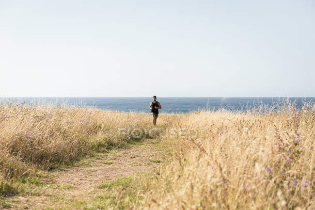 Joggeur masculin musclé courant le long du chemin dans la prairie pendant l'entraînement sur fond de mer en été — Photo de stock