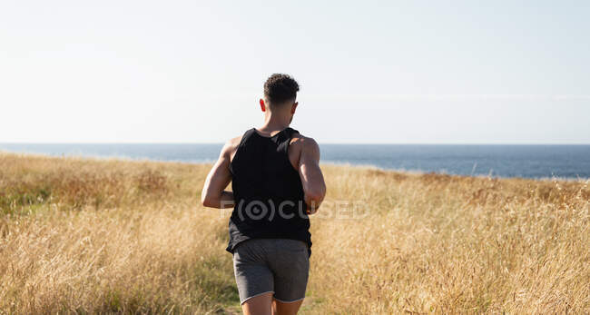 Joggeur masculin musclé courant le long du chemin dans la prairie pendant l'entraînement sur fond de mer en été — Photo de stock