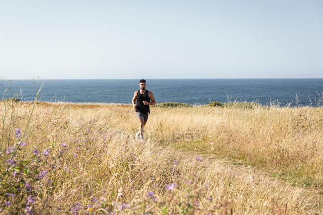 Muscular male jogger running along path in meadow during training on background of sea in summer — Stock Photo