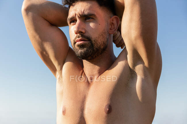 Atleta masculino barbudo sin camisa con cuerpo fuerte de pie con los brazos levantados en el fondo del cielo azul y mirando hacia otro lado - foto de stock