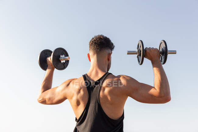 Vista posterior de un guapo atleta masculino con un fuerte torso haciendo ejercicios con pesas mientras está de pie contra el cielo azul en verano - foto de stock