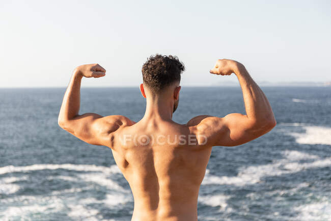 Back view of muscular male bodybuilder with strong naked torso demonstrating biceps while standing near sea in summer — Stock Photo