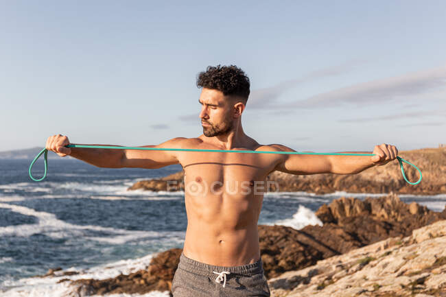 Muscular male bodybuilder with naked torso standing on seashore and doing exercises with resistance band during workout in summer — Stock Photo