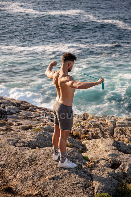 Culturista muscolare maschile con busto nudo in piedi sulla riva del mare e facendo esercizi con banda di resistenza durante l'allenamento in estate — Foto stock