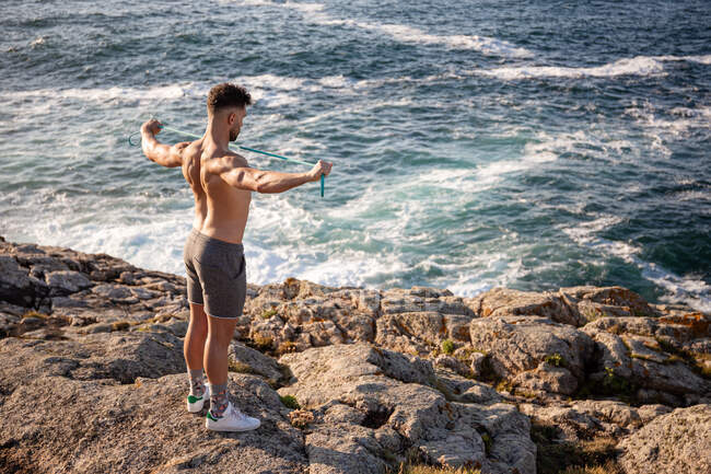 Muscular fisiculturista masculino com tronco nu em pé na praia e fazendo exercícios com banda de resistência durante o treino no verão — Fotografia de Stock