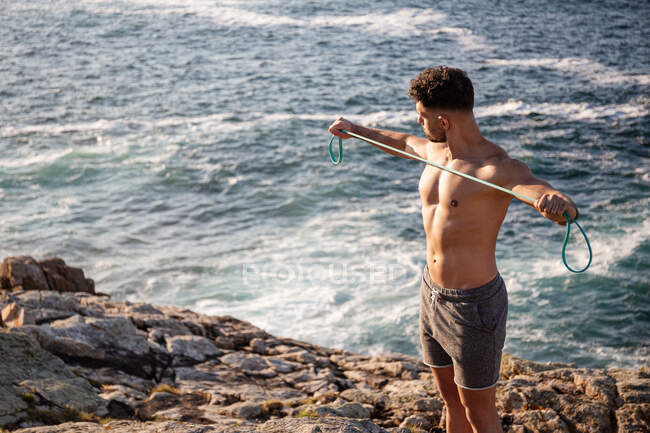 Muscular culturista masculino con torso desnudo parado en la orilla del mar y haciendo ejercicios con banda de resistencia durante el entrenamiento en verano - foto de stock