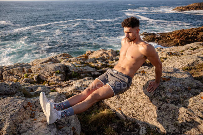 Desde arriba del entrenamiento de atleta masculino sin camisa enfocado en la orilla del mar y haciendo ejercicios de tríceps mientras mira hacia otro lado - foto de stock
