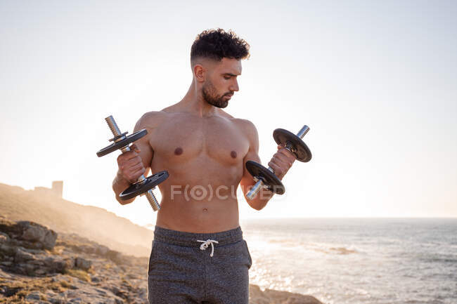 Handsome male athlete with naked torso doing exercises with dumbbells while standing against blue sky in summer — Stock Photo
