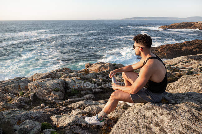 Vista lateral de um macho tranquilo sentado na rocha na costa e admirando a calma paisagem marinha ao pôr-do-sol no verão — Fotografia de Stock