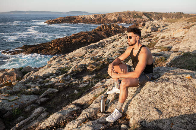 Вид сбоку мирного мужчины, сидящего на скале на берегу и любующегося спокойным мысом на закате летом — стоковое фото