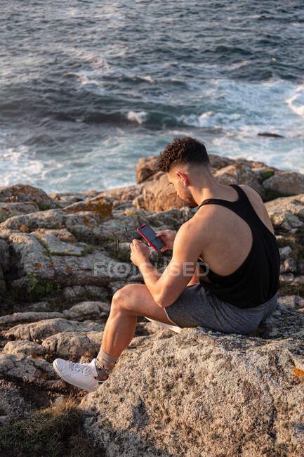 Вид збоку високий кут чоловіка в літньому одязі, що сидить на скелі біля моря і обмін повідомленнями в соціальних мережах через мобільний телефон на заході сонця — стокове фото