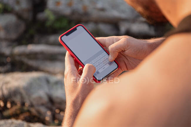Crop high angle of male in summer clothes sitting on rock near sea and messaging on social media via cellphone at sunset — Stock Photo