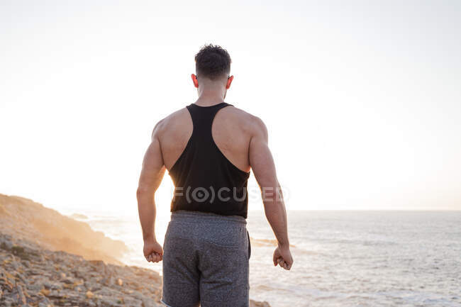 Vista posterior de atleta masculino muscular en ropa deportiva de pie en la playa y disfrutando de la puesta del sol después del entrenamiento en verano - foto de stock