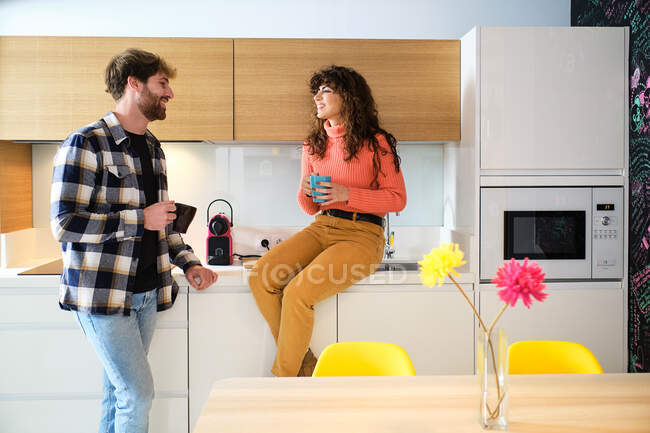 Jovem casal alegre em roupas casuais brilhantes falando na cozinha acolhedora e olhando um para o outro — Fotografia de Stock