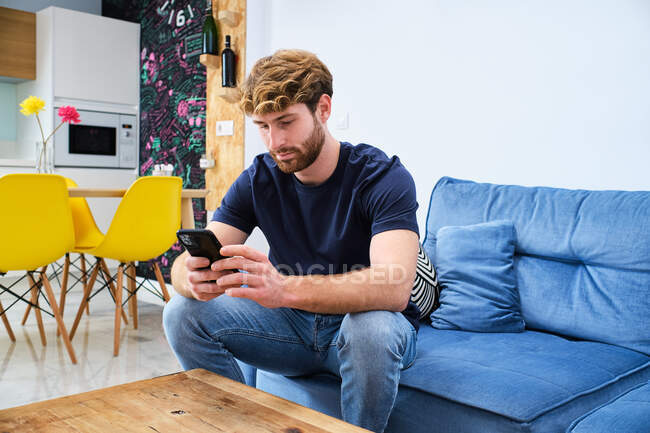 Молодий концентрований чоловік в повсякденному одязі, сидячи на дивані і переглядаючи мобільний телефон в сучасній стильній кімнаті — стокове фото
