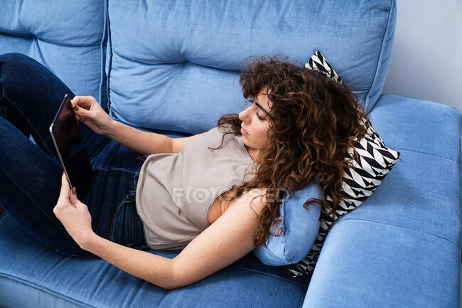 Alto ângulo de jovem do sexo feminino com cabelo encaracolado vestindo roupas casuais deitado no sofá azul e tablet de navegação na sala de estar — Fotografia de Stock