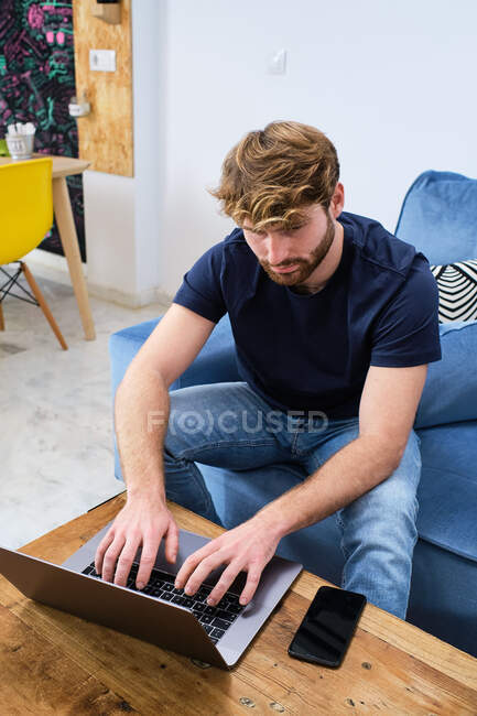 Молодий концентрований чоловік у повсякденному одязі, сидячи на дивані та переглядаючи ноутбук у сучасній стильній кімнаті — стокове фото