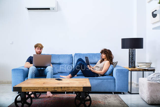 Young woman browsing tablet lying on sofa while man surfing internet on laptop in modern apartment — Stock Photo