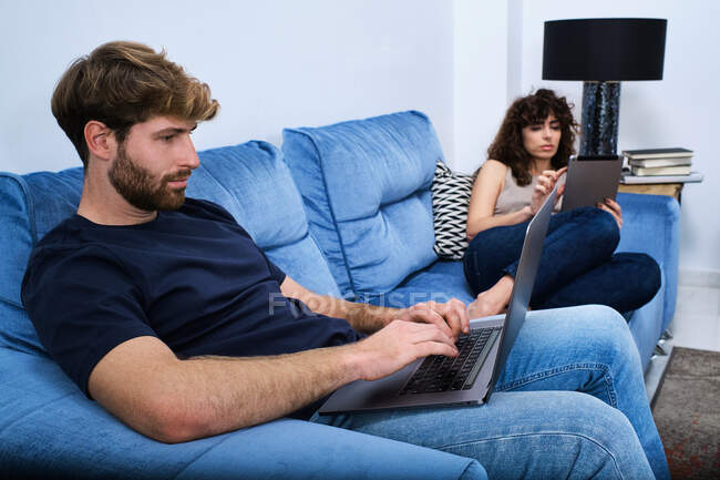 Junge Frau surft Tablet liegend auf Sofa, während Mann in moderner Wohnung auf Laptop im Internet surft — Stockfoto