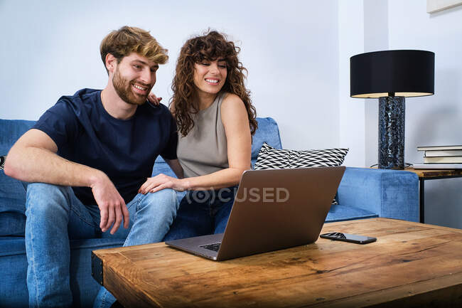 Joven pareja feliz en ropa casual tener conversación en línea en el ordenador portátil sentado en el sofá en la sala de estar - foto de stock