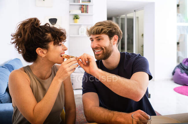 Homem feliz dando fatia de pizza para a namorada enquanto passam o tempo juntos perto do balcão e sofá na sala de luz com prateleiras com livros e elementos decorativos — Fotografia de Stock