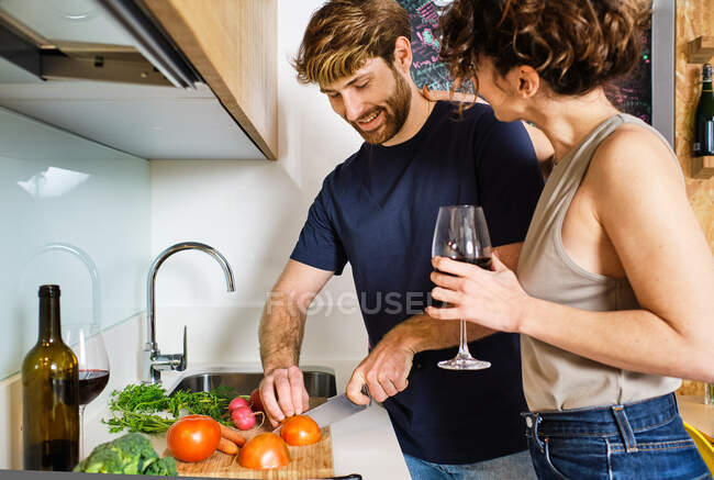 Side view of young couple standing in kitchen with glass of red wine near stove and counter and cutting tomatoes on cutting board with knife near sink and microwave — Stock Photo