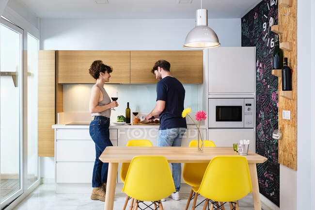 Vista lateral do jovem casal em pé na cozinha com copo de vinho tinto perto do fogão e contador e corte de tomates na placa de corte com faca perto da pia e microondas — Fotografia de Stock