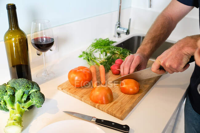 Anonymous man standing in kitchen while cutting tomatoes on cutting board with knife near bottle and glass wine — Stock Photo