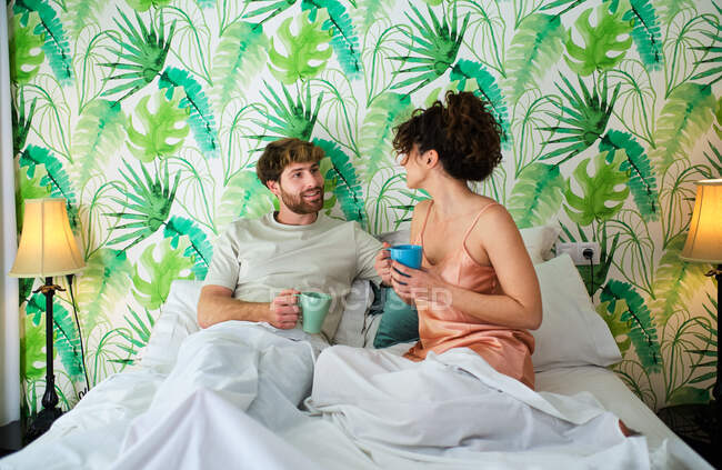 Молода пара в сплячому одязі, насолоджуючись гарячою кавою в ліжку на білій постільній білизні з подушками і ніжно дивлячись один на одного в світлій квартирі — стокове фото