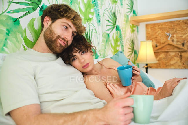 Молодая пара в сонной одежде обнимается, наслаждаясь горячим кофе в постели на белом постельном белье с подушками и глядя на камеру в светлой квартире — стоковое фото