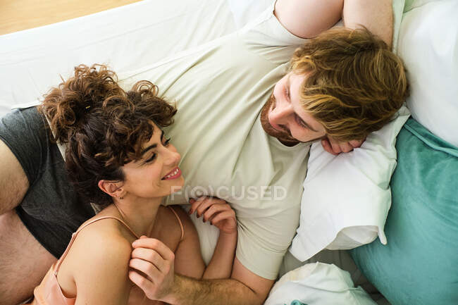 Високий кут молодої пари, що сміється разом у білому ліжку, одягнений у сплячий одяг і лежить один на одного у світлій спальні — стокове фото