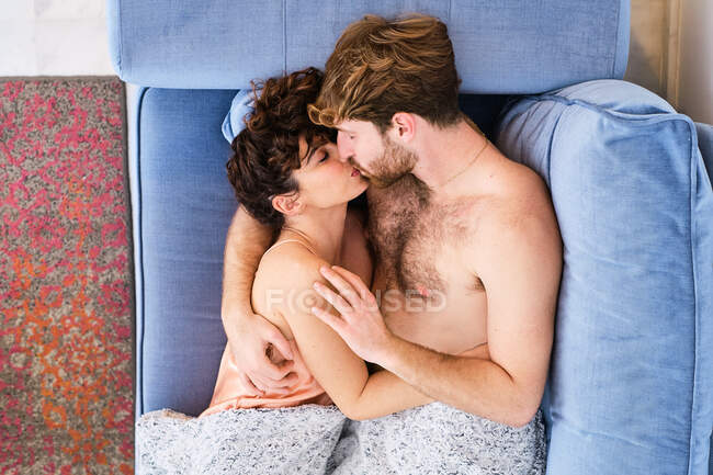 Top view side view of young couple in sleepwear lying on sofa and cuddling while kissing tenderly in light room — Stock Photo