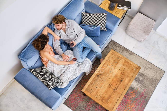 Dall'alto di coppia giovane felice su divano e preparazione per bacio con occhiali con vino rosso in mani e coperta in appartamento leggero — Foto stock