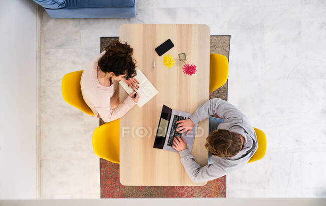 Top view of anonymous female sitting at table on chair and taking notes in notepad with pen near phone and vase with flowers and male using netbook in light room — Stock Photo