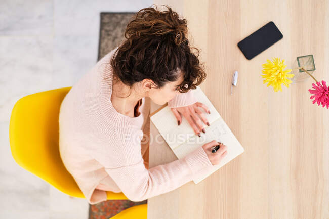 Jeune femme concentrée en tenue décontractée assise à table et écrivant des informations dans un carnet avec stylo près du smartphone et vase en verre avec des fleurs dans la pièce lumineuse — Photo de stock
