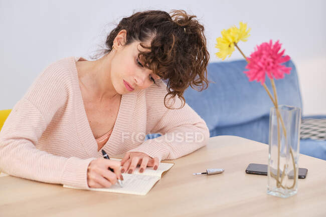Joven dama concentrada en traje casual sentada en la mesa y escribiendo información en cuaderno con pluma cerca de teléfono inteligente y jarrón de cristal con flores cerca del sofá en la sala de luz - foto de stock