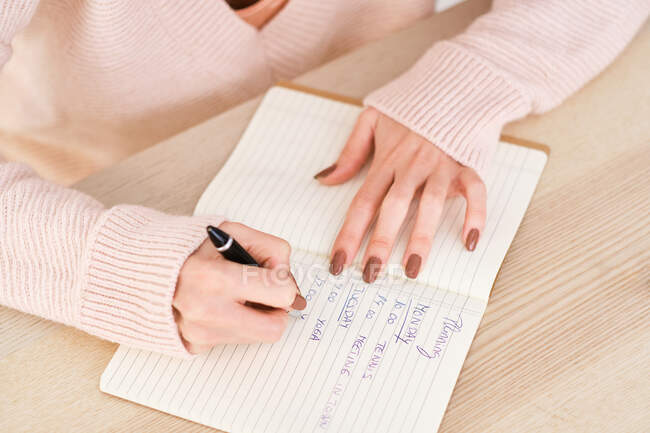 Высокий угол безликой женщины в повседневном наряде, сидящей за деревянным столом и пишущей план недели в тетрадке с ручкой в светлой комнате — стоковое фото