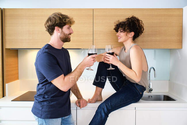 Vista laterale di giovane coppia sorridente in abiti casual con bicchieri di vino rosso tifo in cucina luminosa vicino armadi e lavandino con rubinetto e guardarsi l'un l'altro — Foto stock