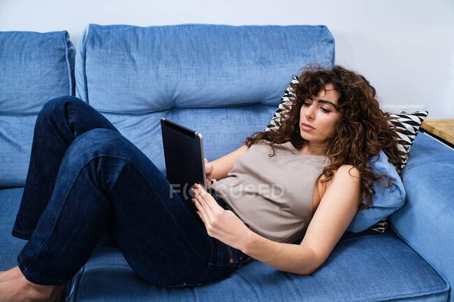 Високий кут молодої жінки з кучерявим волоссям у повсякденному одязі, що лежить на синьому дивані та планшеті у вітальні — стокове фото