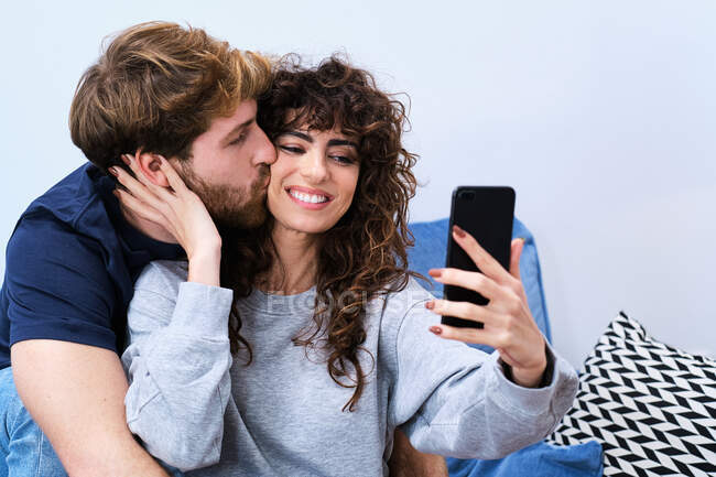 Молодой бородатый мужчина целует улыбающуюся женщину в повседневной одежде, делая селфи на мобильный телефон — стоковое фото