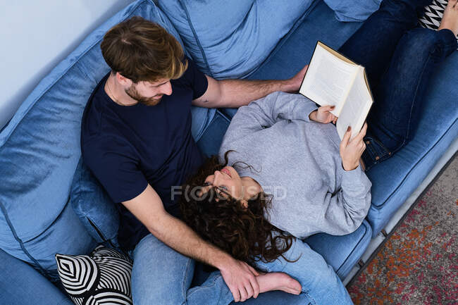 De arriba la mujer joven en la ropa casual acostado en el sofá y el libro de lectura poniendo la cabeza sobre las rodillas del novio - foto de stock