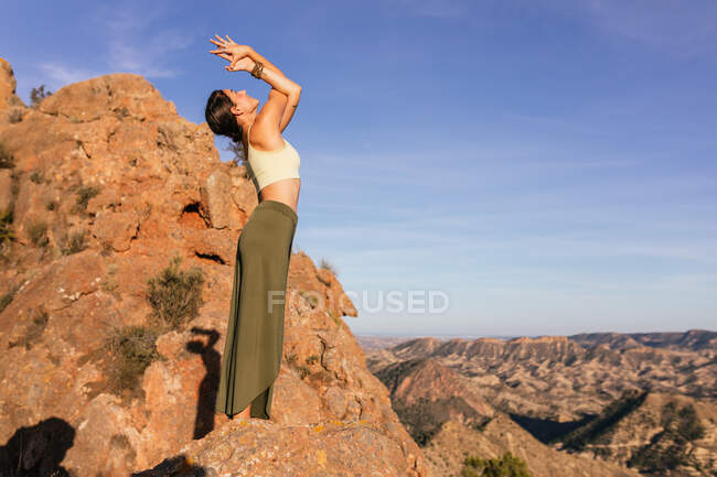 Vista lateral de la hembra delgada en traje casual que realiza la montaña con los brazos hacia arriba y la postura Backbend en la pendiente de la montaña rocosa - foto de stock