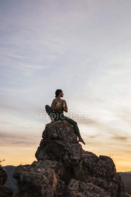 Low angle of calm female performing yoga exercise while practicing meditation on rocky ground at bright sunset — Stock Photo