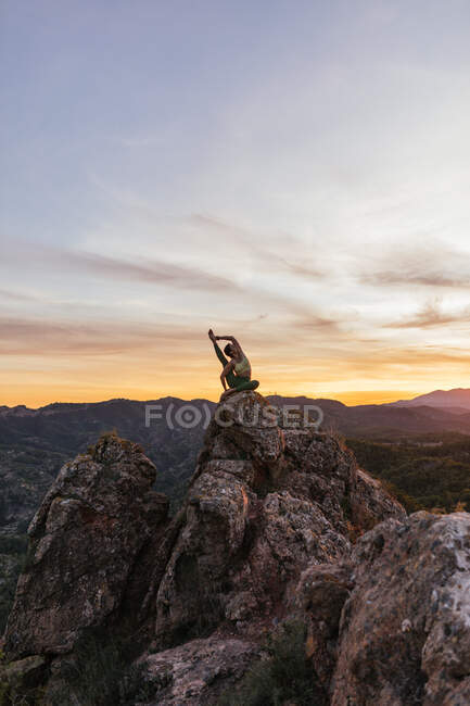 Mujer flexible en ropa deportiva realizando pose de garza en la cima de una colina rocosa mientras practica yoga - foto de stock