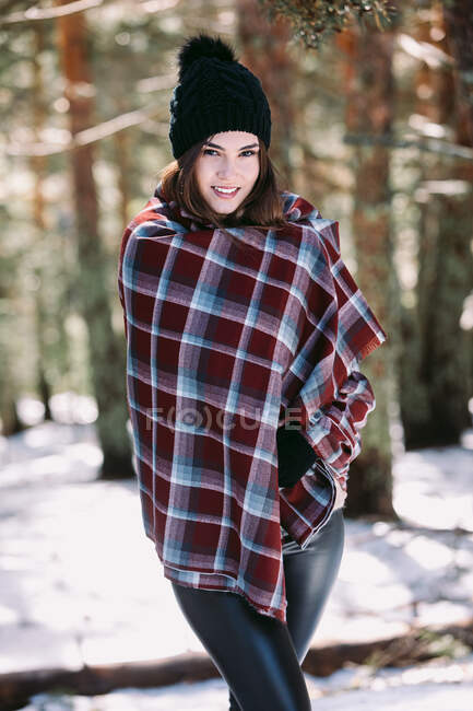 Content female wrapped in warm plaid standing in snowy woods and looking at camera — Stock Photo