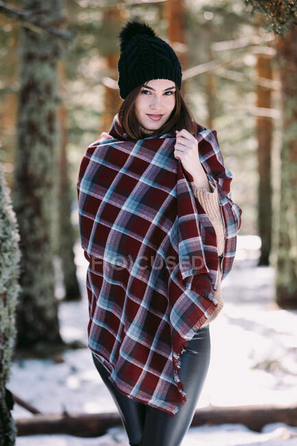 Zufriedene, in warmes Plaid gehüllte Frau steht in verschneiten Wäldern und blickt in die Kamera — Stockfoto