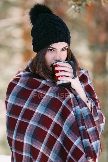 Жінка, загорнута в теплий плед, сидить на стовбурі дерева в засніженому зимовому лісі і насолоджується гарячим напоєм з чашки — стокове фото